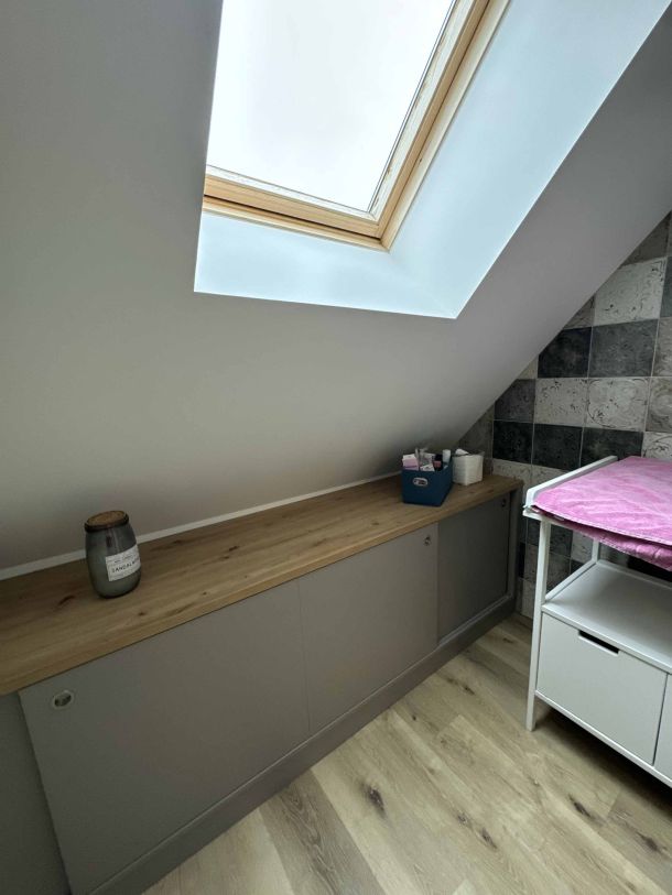 Meuble bas sur mesure en finition grise et bois clair, placé sous une fenêtre de toit avec un sol en parquet, complété par une décoration minimaliste et un mur texturé en carrelage.